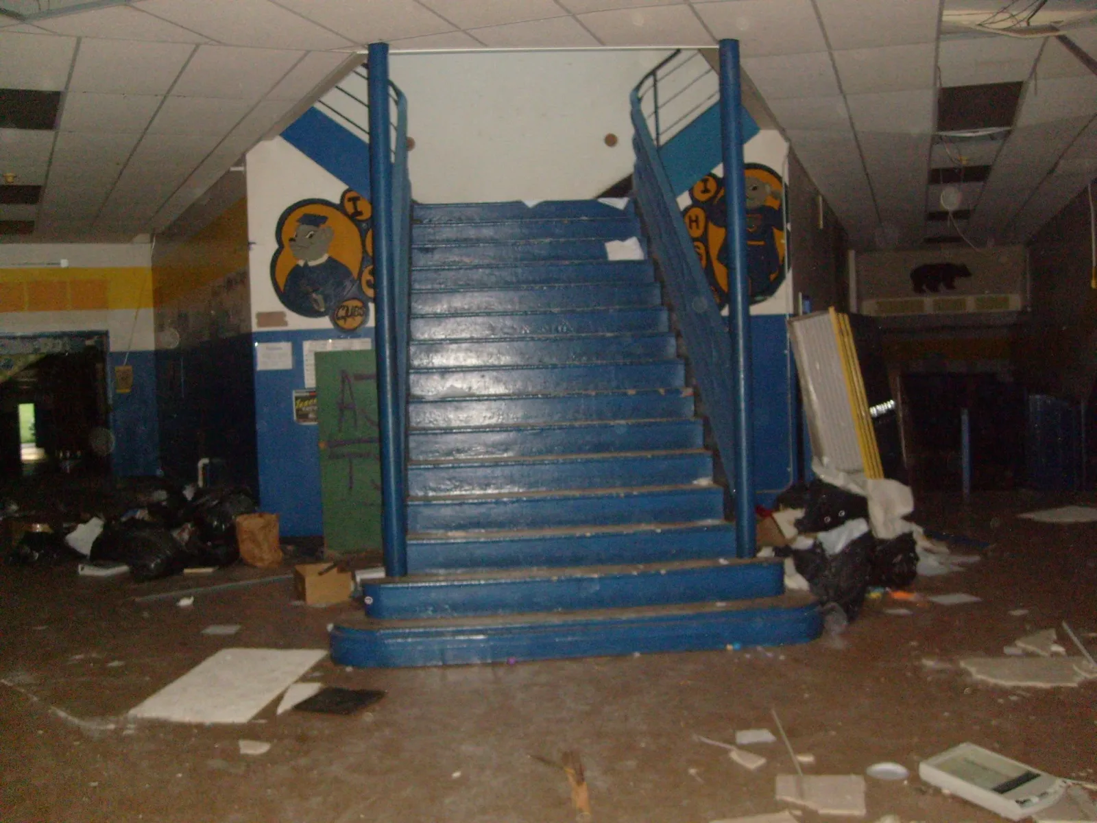 Double-sided blue stairway in a school lobby with murals on either side, a green locker with graffiti to the left, scattered debris and missing ceiling tiles.